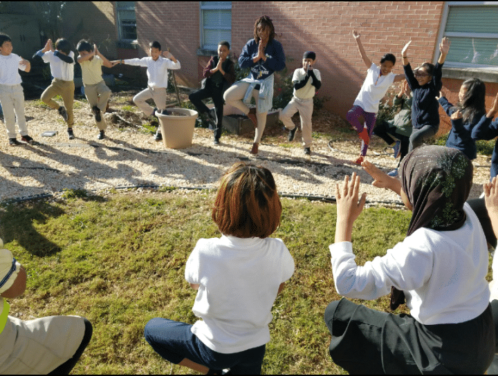 FoodCorps alum Zashe Cockett-Demings (GA '18) stands in a circle of students outdoors; some are balancing on one foot, others are holding their hands out. Movement and garden exposure in lessons encourages positive mental health among students.